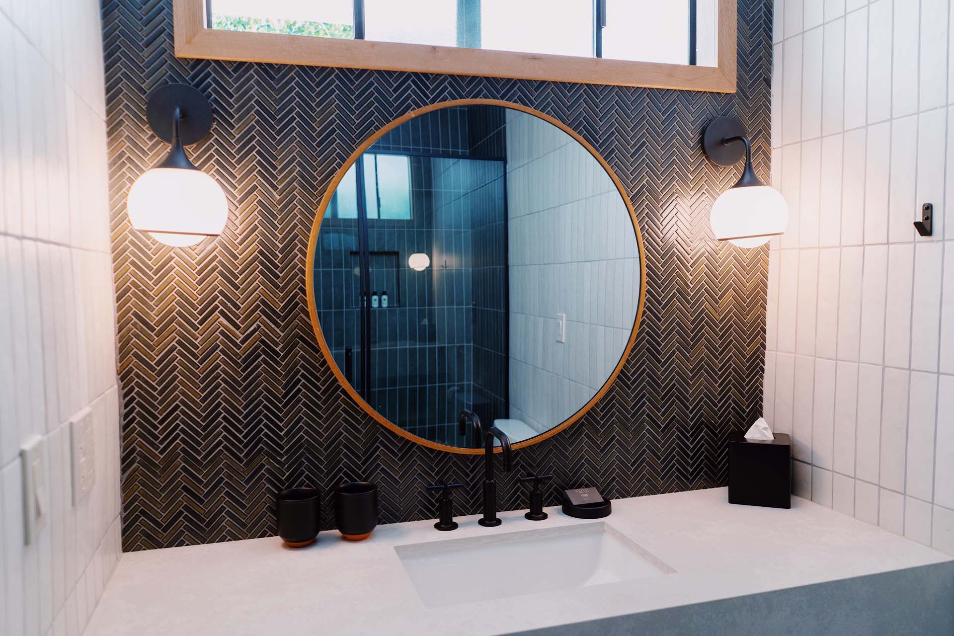 A modern bathroom sink area with a round mirror, herringbone tile backsplash, and black and white decor.