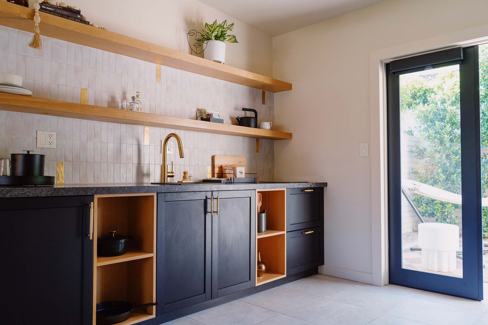 A kitchenette with black cabinets, wooden shelves, and a sink, with a door leading to an outdoor area.