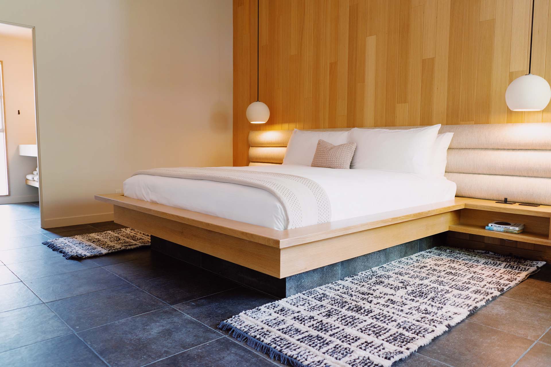 A modern minimalist hotel room with a platform bed, wooden headboard, pendant lights, tiled floor, and area rug.