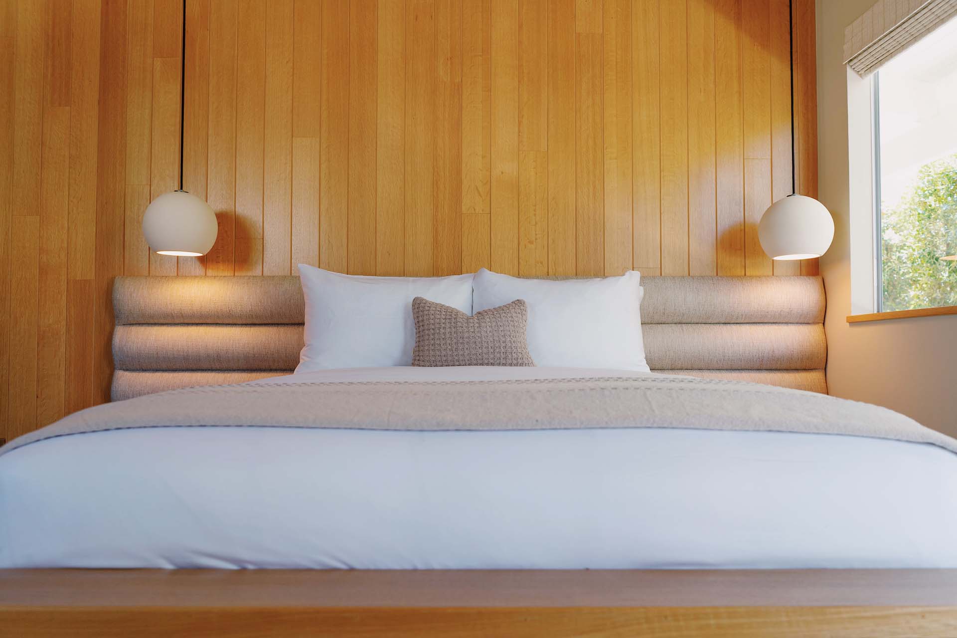 A neatly made bed with white linens and pillows, with a wood-paneled headboard and pendant lights on either side.