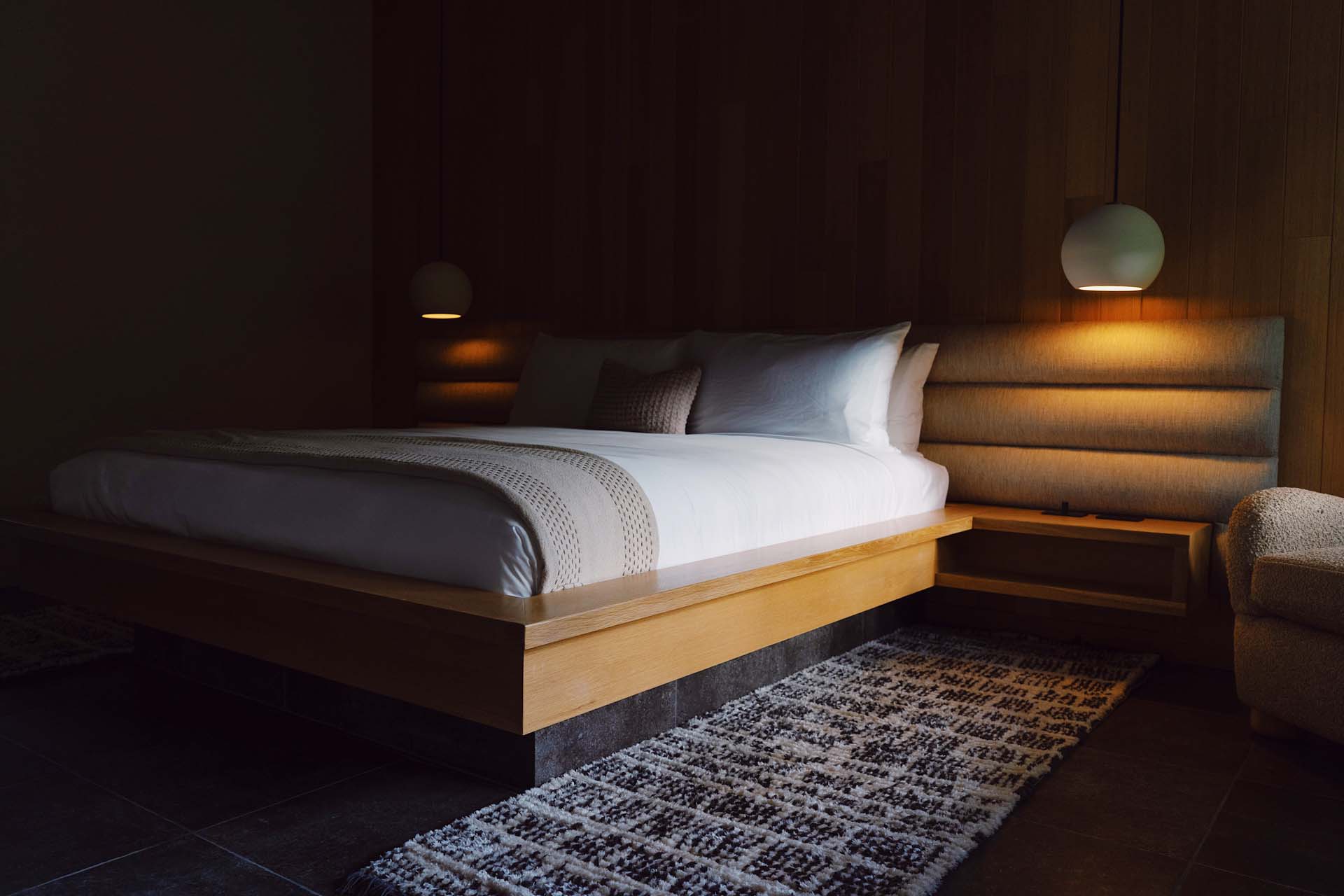 A neatly made bed with white linens and pillows, a nightstand with a lamp, and a chair in a dimly lit room.