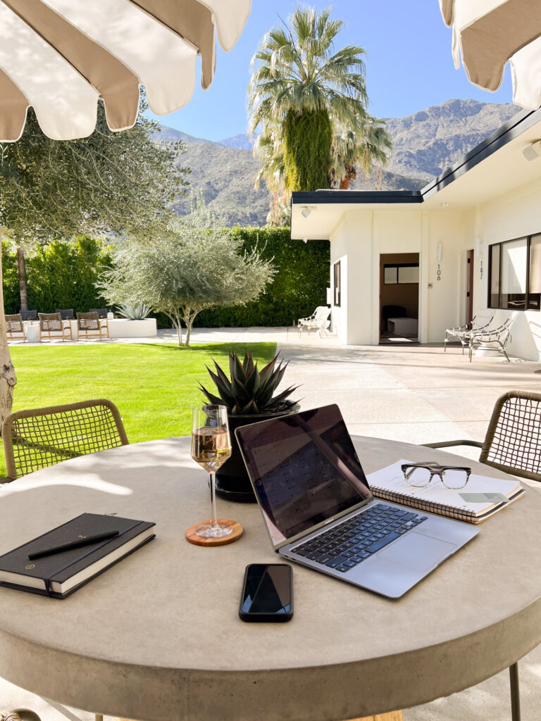 Remote worker with laptop at shaded Palm Springs boutique hotel courtyard table with pool in background